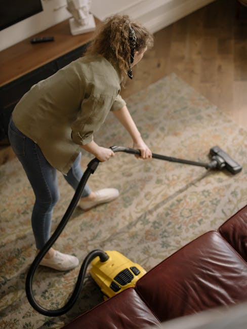 A professional cleaner from Carpet Cleaners SW3 equipped with a vacuum hose and wearing protective clothing, gloves, and shoe covers, performing deep surface cleaning on a light-colored, plush carpet 