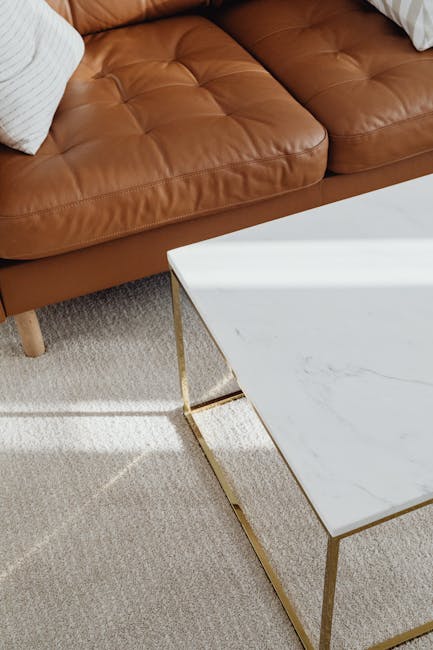 A close-up view of a modern living room corner featuring a brown leather sofa with tufted cushion details and a white textured pillow. Next to the sofa is a sleek, rectangular marble-topped coffee table with thin gold legs, situated on a light beige, plush carpet. The room is well-lit, highlighting the clean, polished surfaces and overall tidy appearance. This setting exemplifies domestic surface cleaning and maintenance, indicative of professional services offered by Carpet Cleaners SW3 for deep cleaning and sanitisation of home interiors.