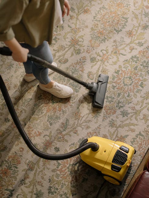 A person using a yellow vacuum cleaner with a black hose attachment to deep clean a traditional patterned rug in a residential living room. The rug features a floral design in muted tones of beige, green, and orange, and covers a wooden floor. The individual's legs are visible, wearing light-colored sneakers and casual clothing, with the person holding the vacuum's hose nozzle firmly on the carpet. Natural light illuminates the space, highlighting the cleanliness and well-maintained appearance of the rug and surrounding area. Carpet Cleaners SW3 specializes in surface cleaning and sanitisation, ensuring homes like this are hygienic and visually fresh.