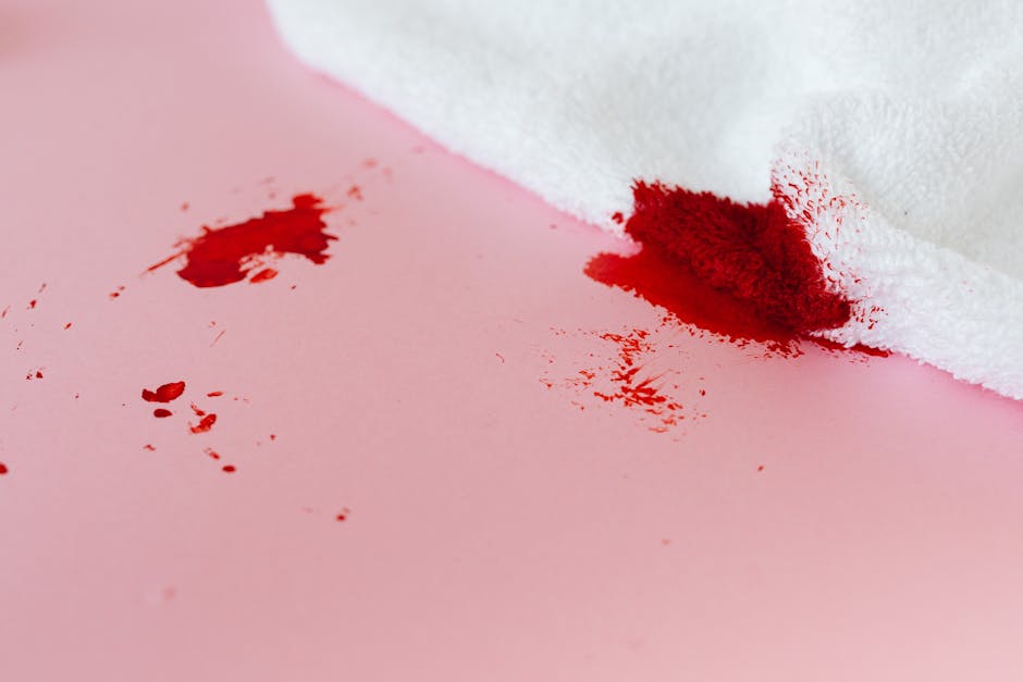 Close-up image showing a white cloth partially covering bright red wine spill on a light pink surface, likely a table or countertop. The spill appears to be recent, with some streaks and scattered dri